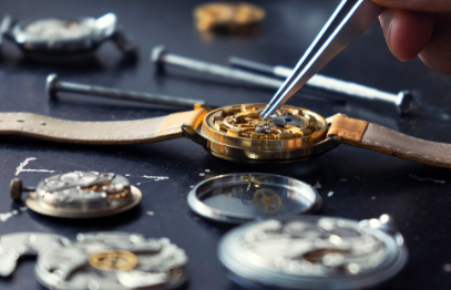 Mano utilizando pinzas para reparar el mecanismo abierto de un reloj rodeado de piezas y herramientas de relojería.